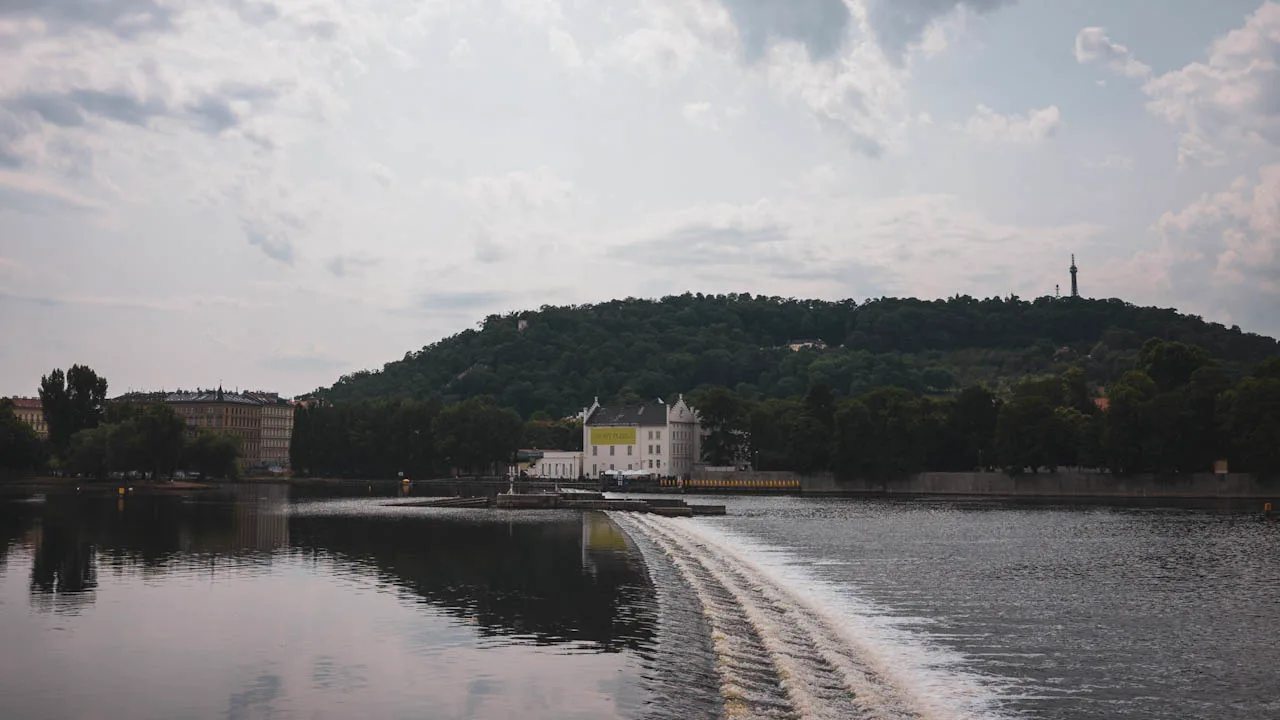 The Vltava weir under a cloudy sky, a wooded hill with a lookout tower behind